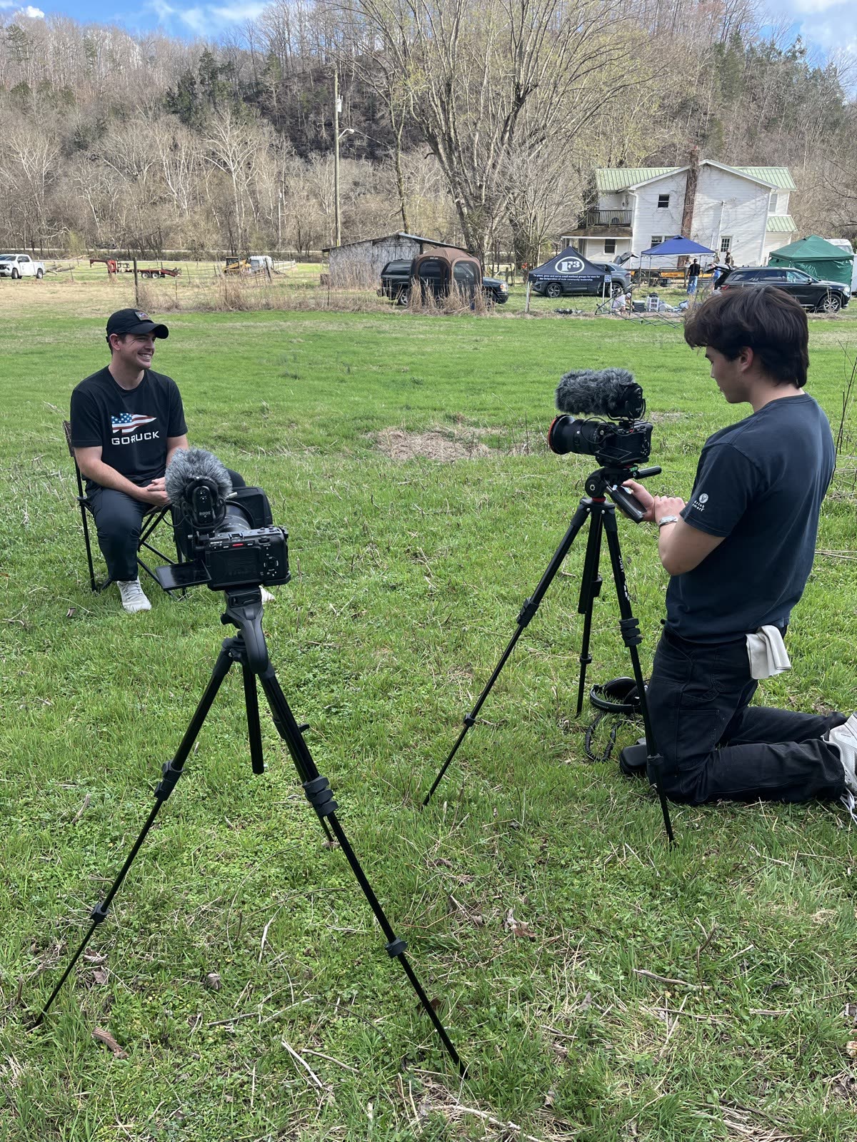 A two-camera interview in the field, farmhouse and race canopies in the distance.