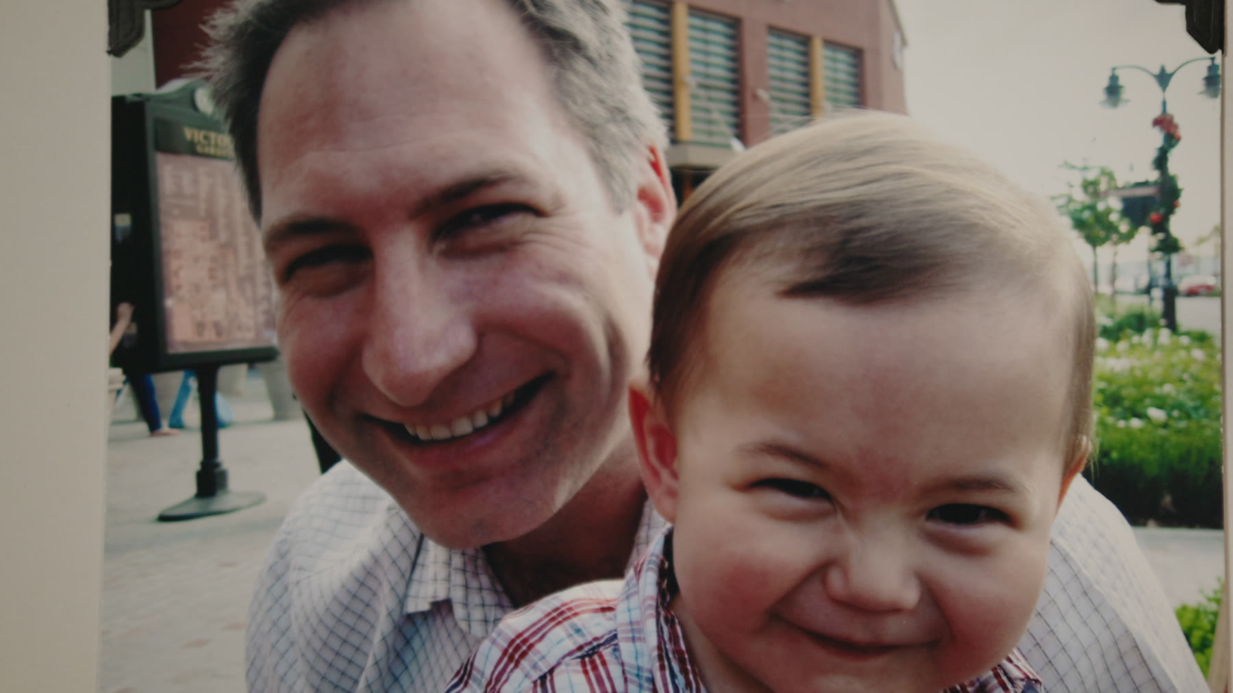 An archival snapshot of a man holding a laughing baby outdoors in a town square.